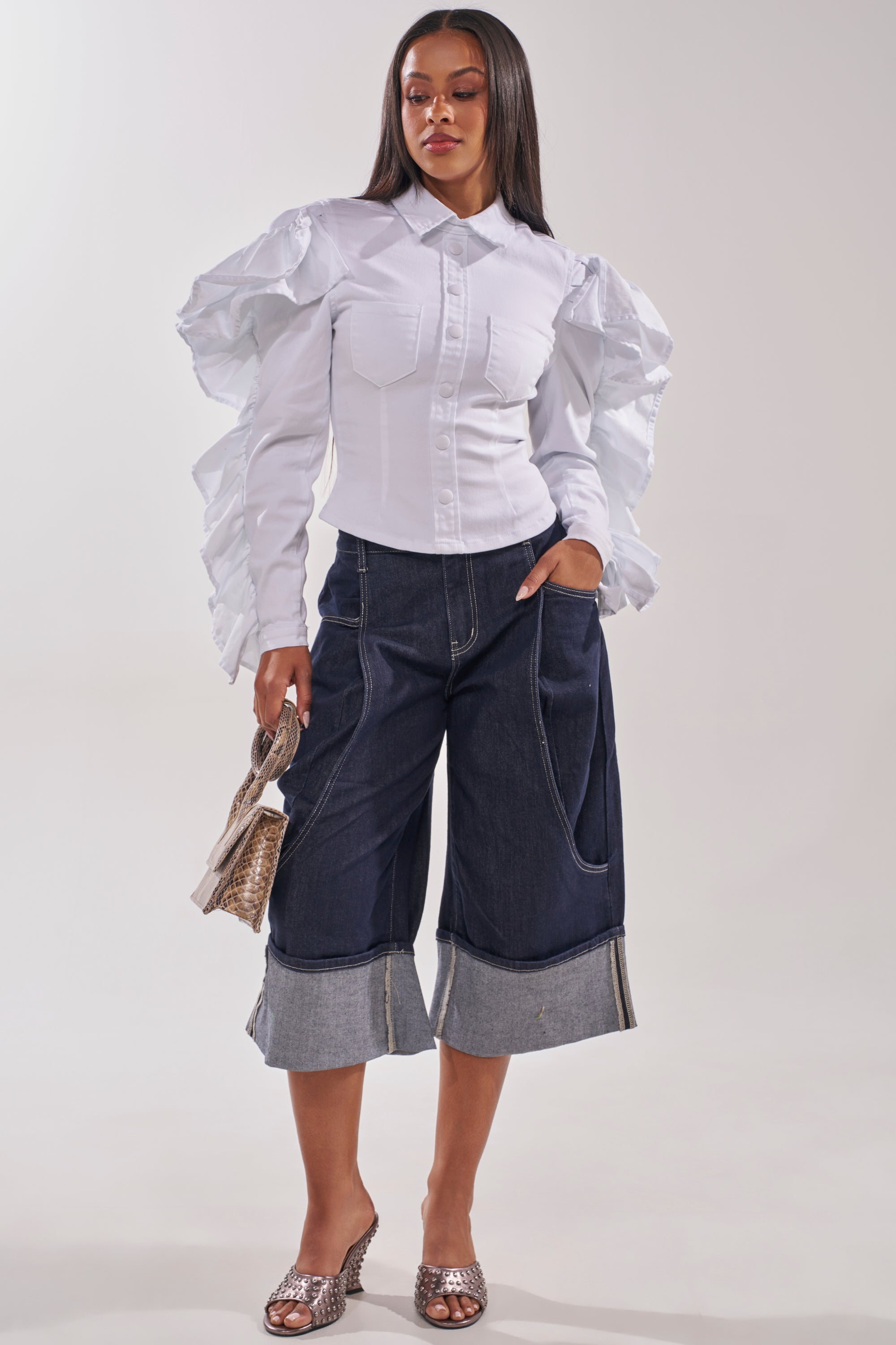 A woman wears a white ruffled sleeve shirt, the COME WITH ME DENIM BERMUDA SHORT in dark wash stretch cotton, sparkly shoes, and carries a small handbag. 6
