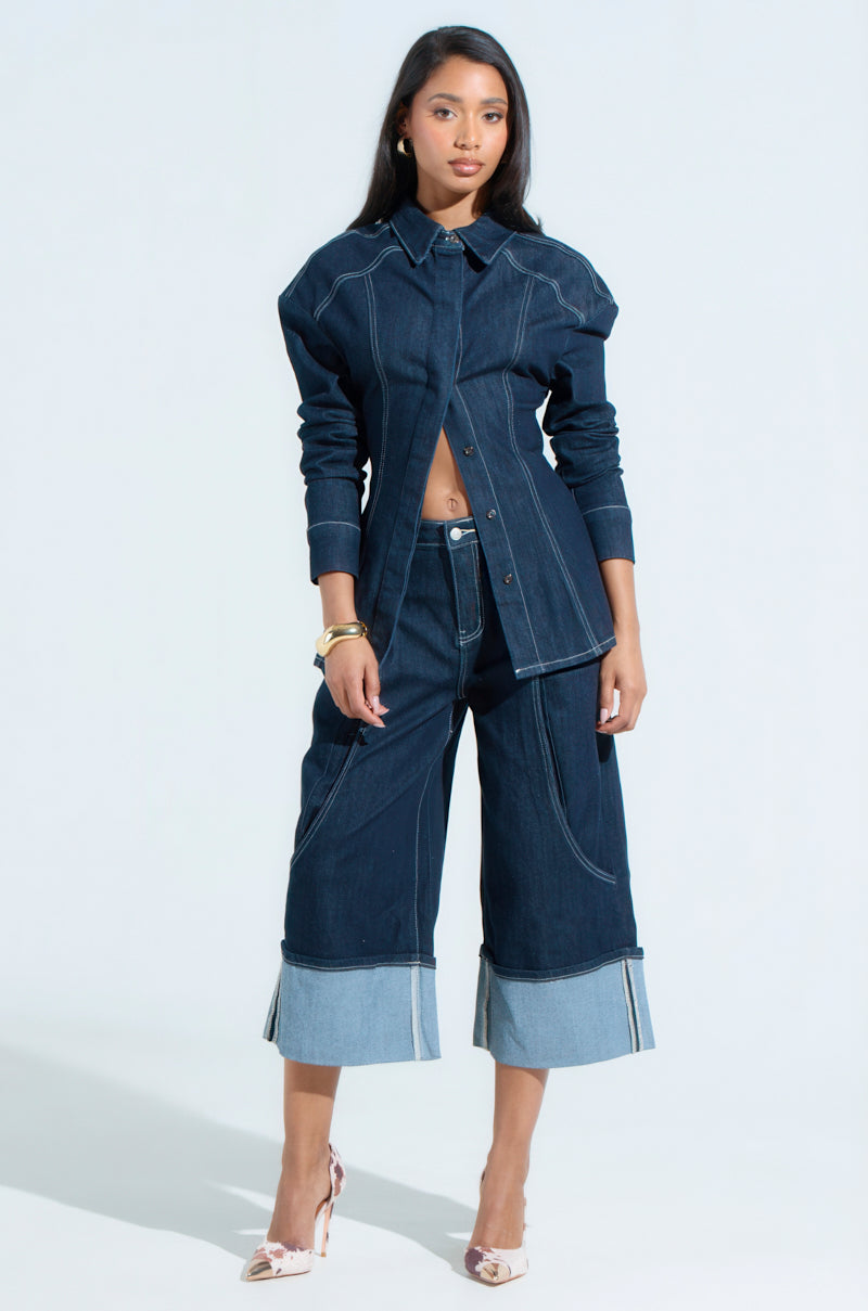 A woman poses against a light background wearing the COME WITH ME DENIM BERMUDA SHORT and a dark denim jacket. She accessorizes with bracelets and high-heeled shoes, her straight dark hair mirroring her outfit's structure, as she looks confidently at the camera.