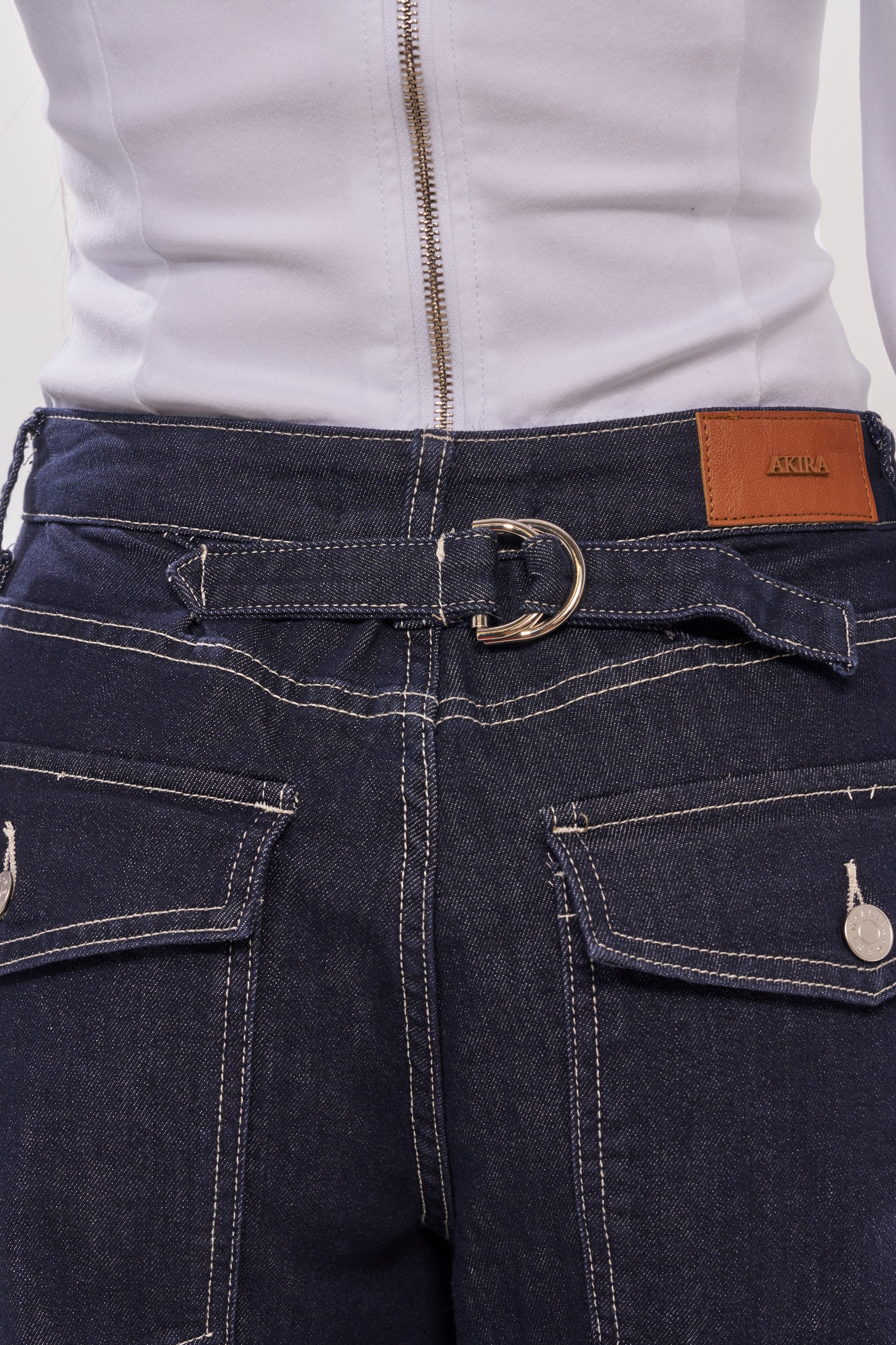 Close-up of the COME WITH ME DENIM BERMUDA SHORT in dark wash, featuring white stitching, large pockets, a matching denim belt with metal ring buckle, a brown leather patch on the waistband, and paired with a white zippered top—ideal for festivals. 8