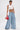 A woman wearing a white sleeveless crop top with colorful flowers, THE BEST ONE YET WIDE LEG DENIM jeans, and holding a small red handbag. She has tattoos, slicked-back hair, and poses against a plain white background.