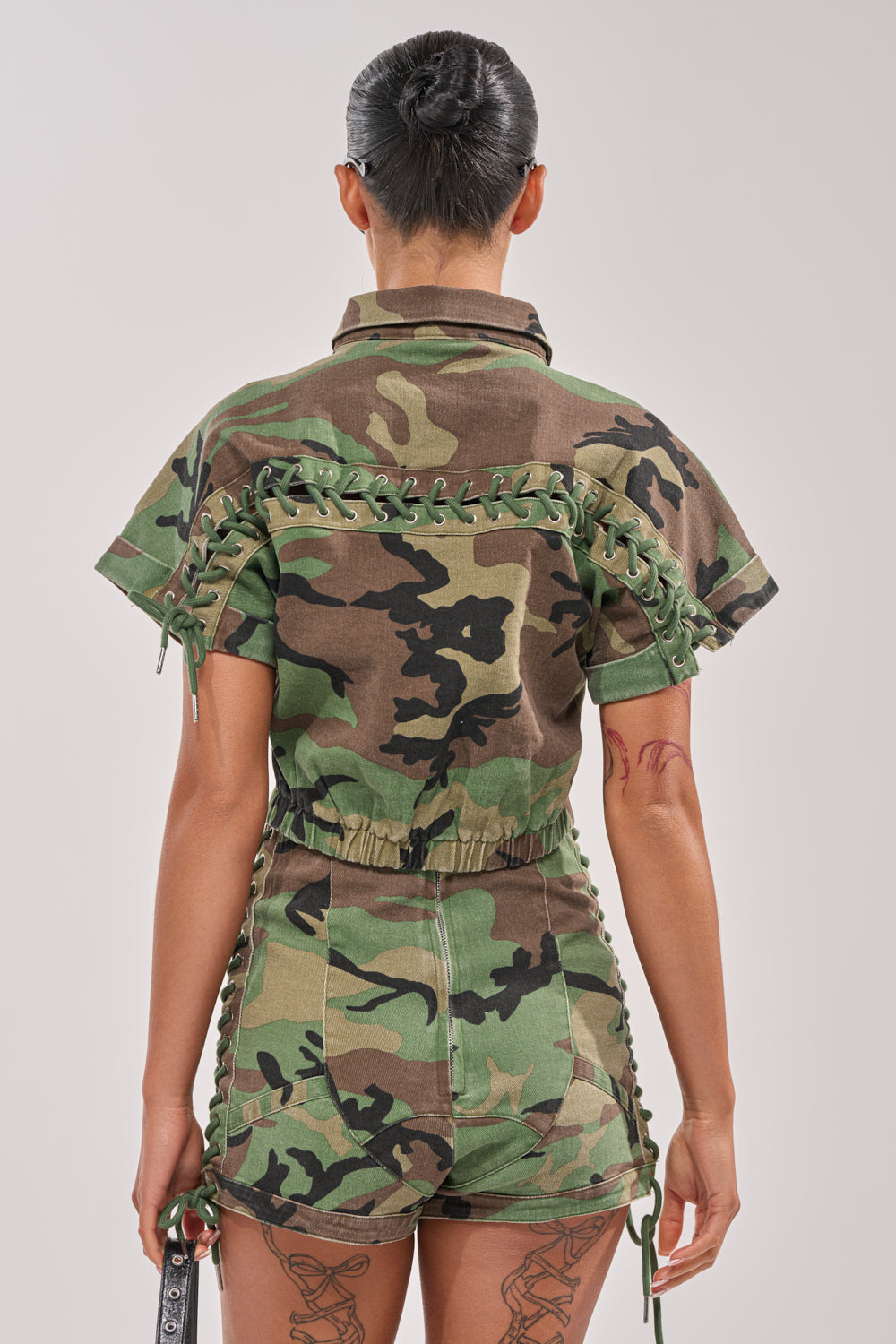 A person with short, dark hair is seen from the back wearing the I MEAN BUSINESS CAMO BUTTON DOWN, styled street-style to showcase their tattoos on both arms and legs. 4