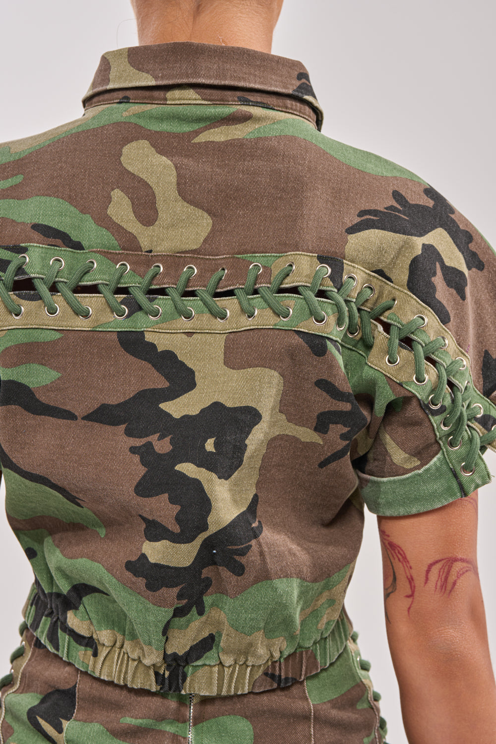 The I MEAN BUSINESS CAMO BUTTON DOWN features green lacing detail across the back and sleeve, styled here on a person with a short haircut and visible red-line tattoo, giving this street-style shirt an edgy vibe. 8