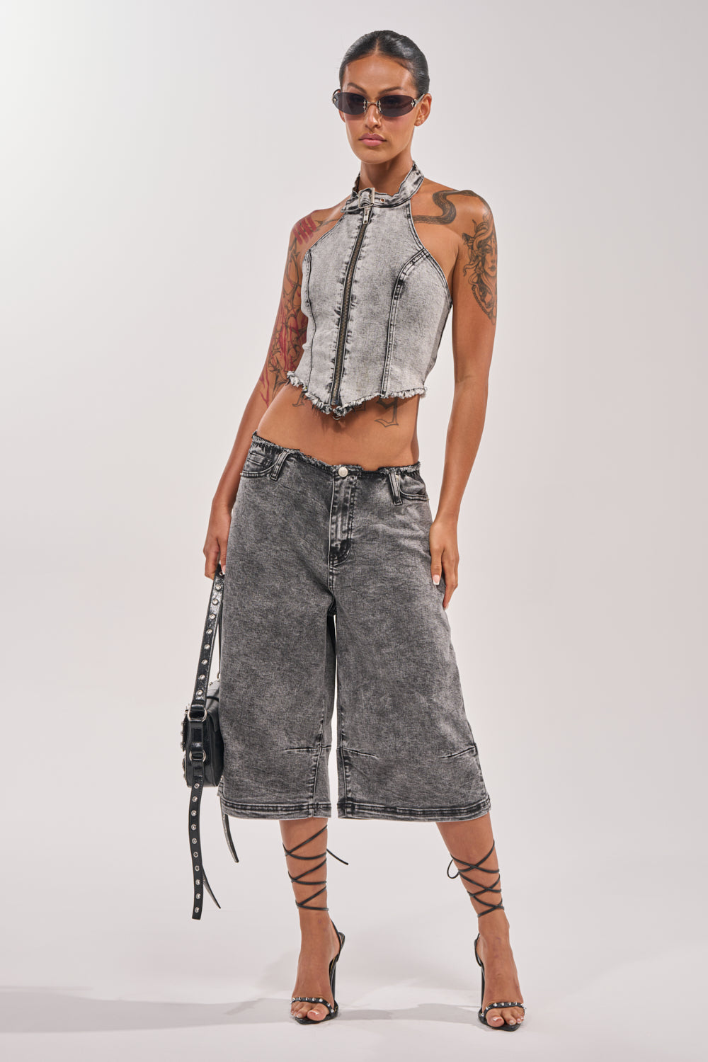 A woman in sunglasses, a sleeveless zip-up gray denim crop top, BREAK UP WITH HIM DENIM SHORTS IN DARK WASH, strappy black heels, and holding a studded black handbag poses confidently against a plain light background. 0