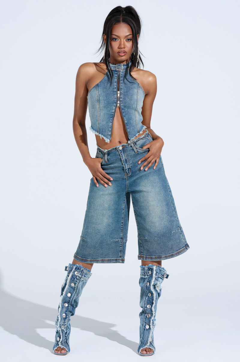 Confidently posing against a light background, the person wears a relaxed silhouette featuring sleeveless denim halter top, BREAK UP WITH HIM DENIM SHORTS in high-rise knee-length style, and knee-high denim boots with embellishments, showcasing diverse textures and patterns.