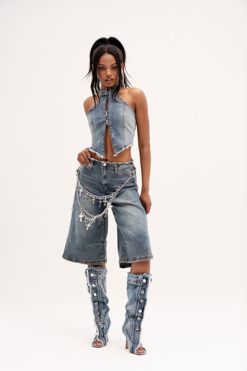 A person wears a denim strapless top with a front zipper and the "BREAK UP WITH HIM DENIM SHORTS," knee-length and high-rise. They accessorize with a chain belt and denim boots adorned with white buttons, against a plain white background that highlights the relaxed silhouette.