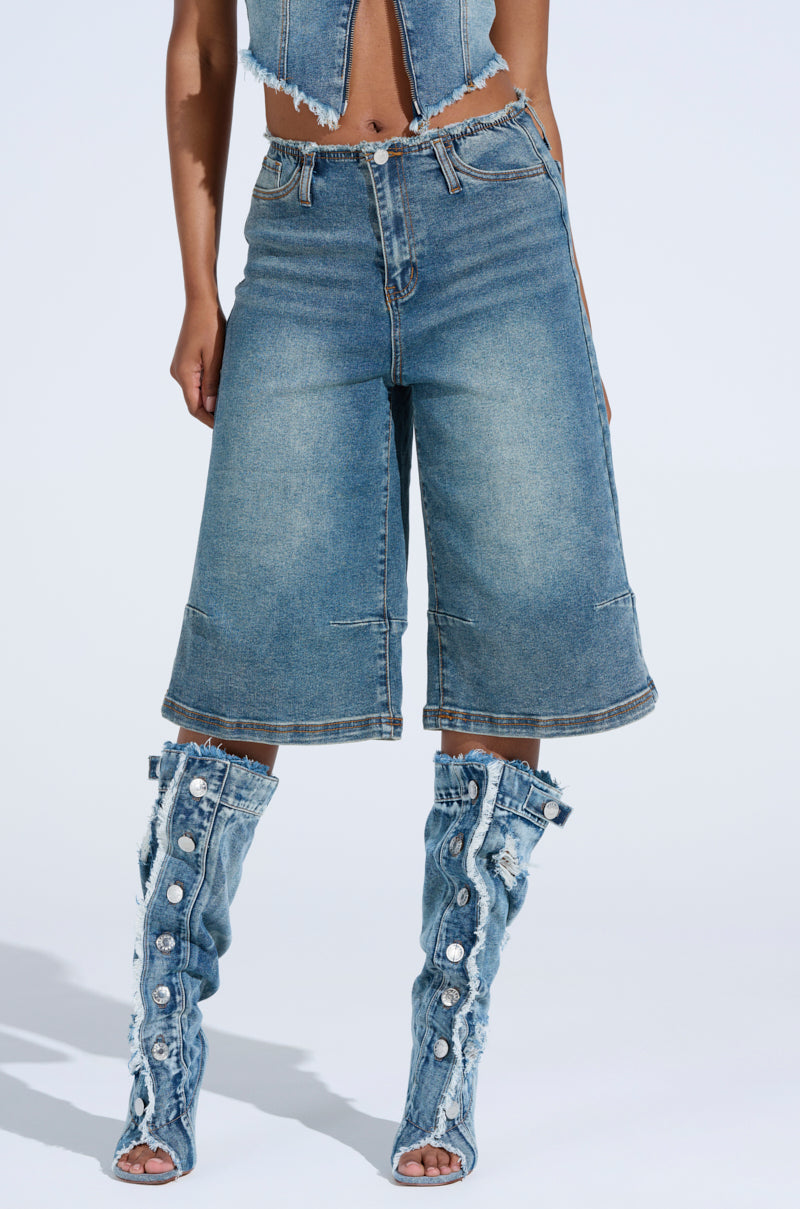A person is dressed in a denim outfit featuring the BREAK UP WITH HIM DENIM SHORTS, which boast a high-rise, knee-length, relaxed silhouette. They're also wearing denim boots with button details and frayed edges. The setting is an off-white background.