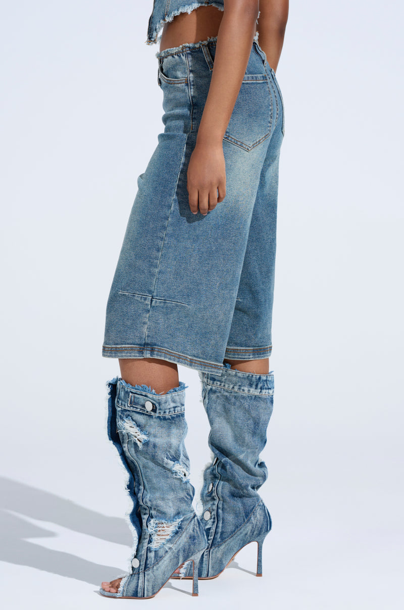 Against a light background, someone showcases the BREAK UP WITH HIM DENIM SHORTS featuring a relaxed silhouette and high-rise fit. Paired with high-heeled denim boots, the faded shorts highlight contrast stitching as the person's hand rests by their side.