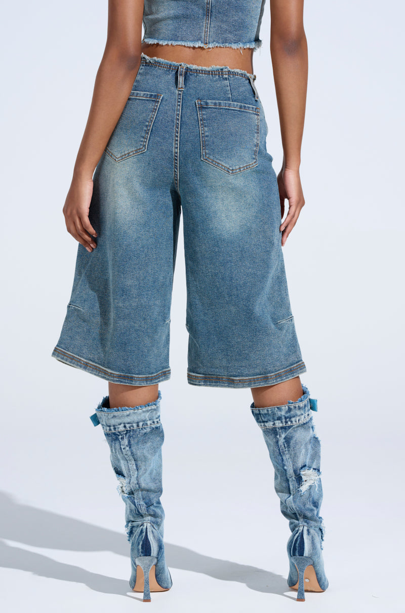 Someone is wearing the BREAK UP WITH HIM DENIM SHORTS featuring a relaxed silhouette. They pair it with a matching top and unique cut-out denim boots tied at the back, standing against a clear, light blue background.