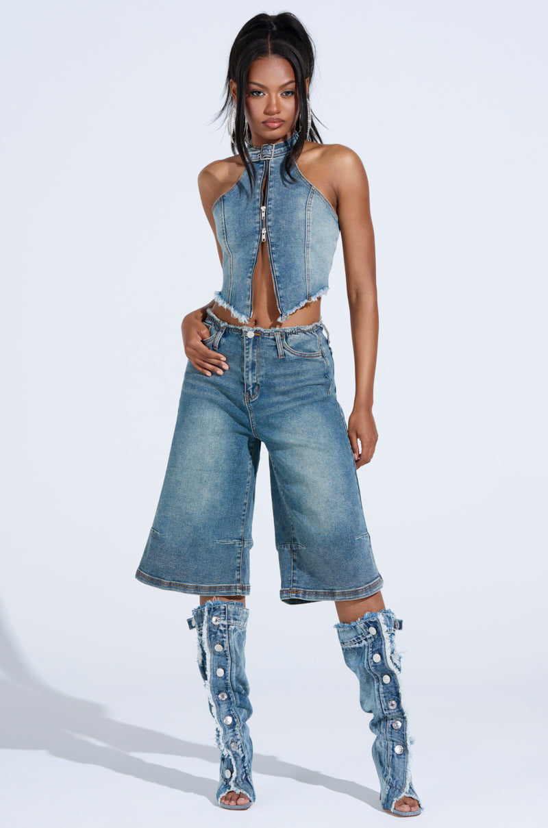 Against a plain background, a person confidently poses in a denim outfit, featuring the "BREAK UP WITH HIM DENIM SHORTS," a halter-style denim top, and knee-high boots with button details. Their hair is styled long and straight.