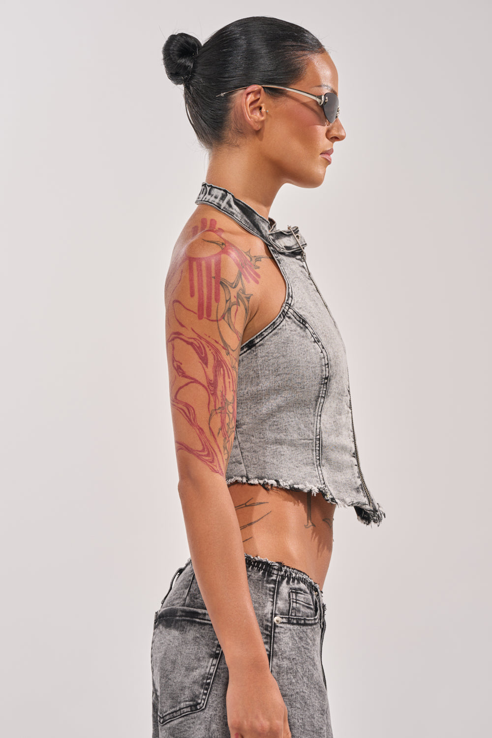 A woman with slicked-back hair, sunglasses, and arm tattoos wears the BREAK UP WITH HIM DENIM TOP—a sleeveless, frayed washed grey halter—paired with matching pants, standing in profile against a plain light background. 2