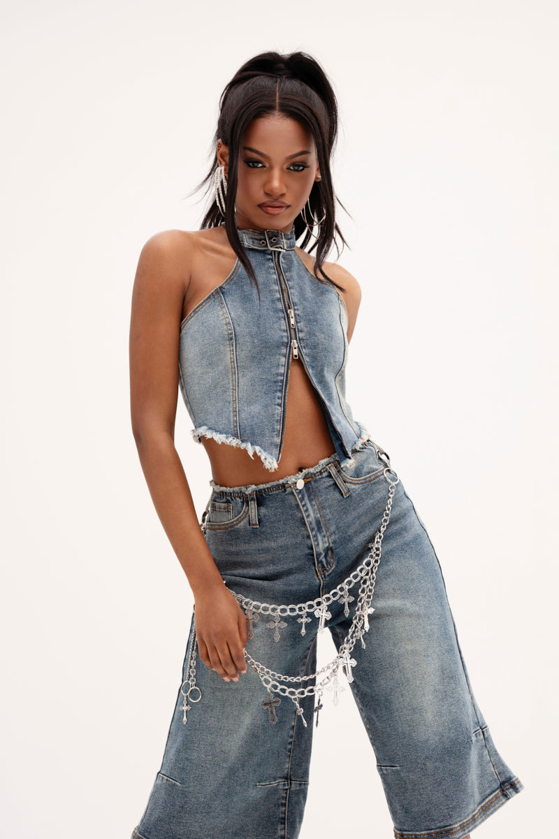 Sporting a high ponytail and silver chain accessories, a person models the BREAK UP WITH HIM DENIM HALTER TOP paired with wide-legged, medium-wash jeans. The outfit features frayed edges and stands out against a plain background.