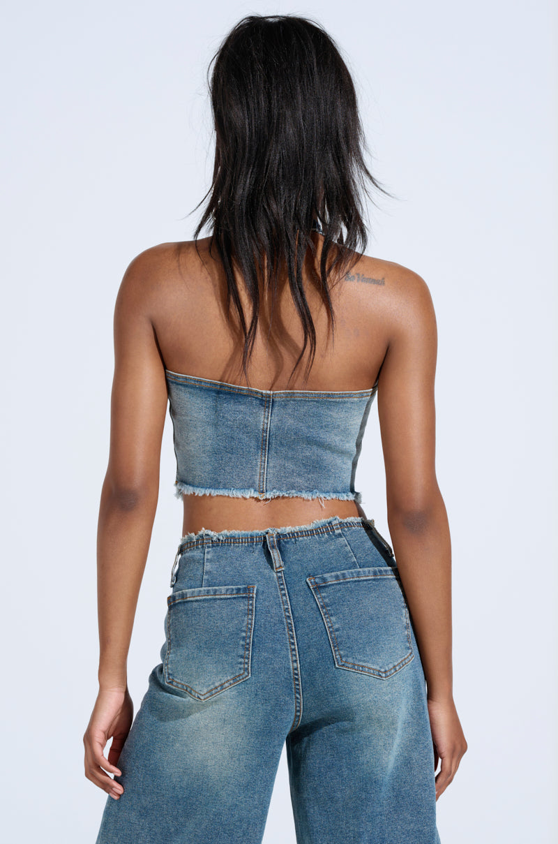 A person with long hair is wearing the AKIRA Label's BREAK UP WITH HIM DENIM HALTER TOP and high-waisted jeans, facing away from the camera against a light background.