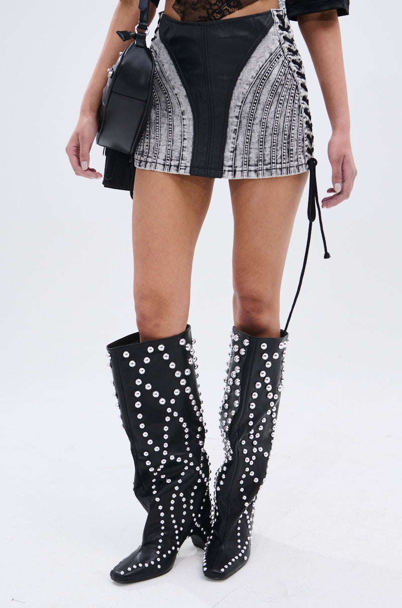 A person wears the TELL ME LIES MINI SKIRT, a black faux leather side-lace-up top, tall black studded boots, and holds a black handbag against a plain light background.