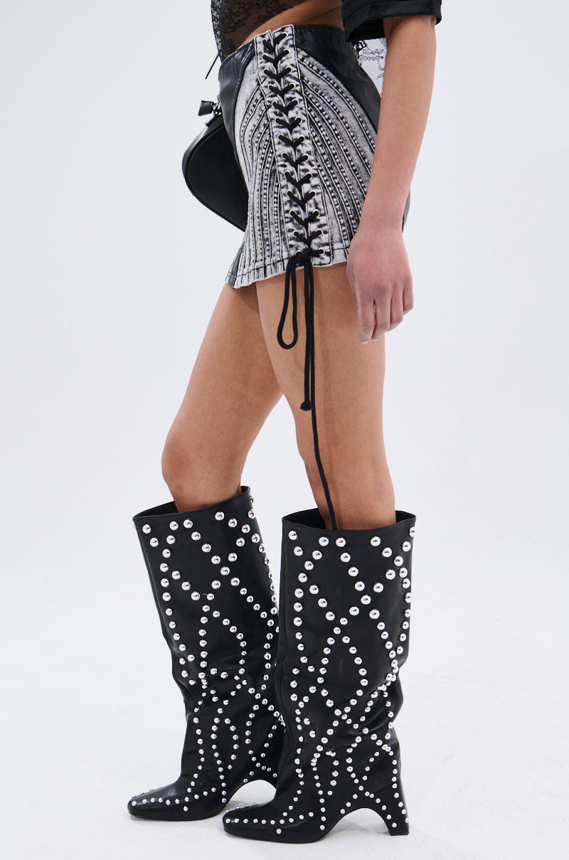 A person stands sideways against a plain light background, wearing black knee-high boots with white studs, the TELL ME LIES MINI SKIRT featuring lace-up side details, and holding a black bag.