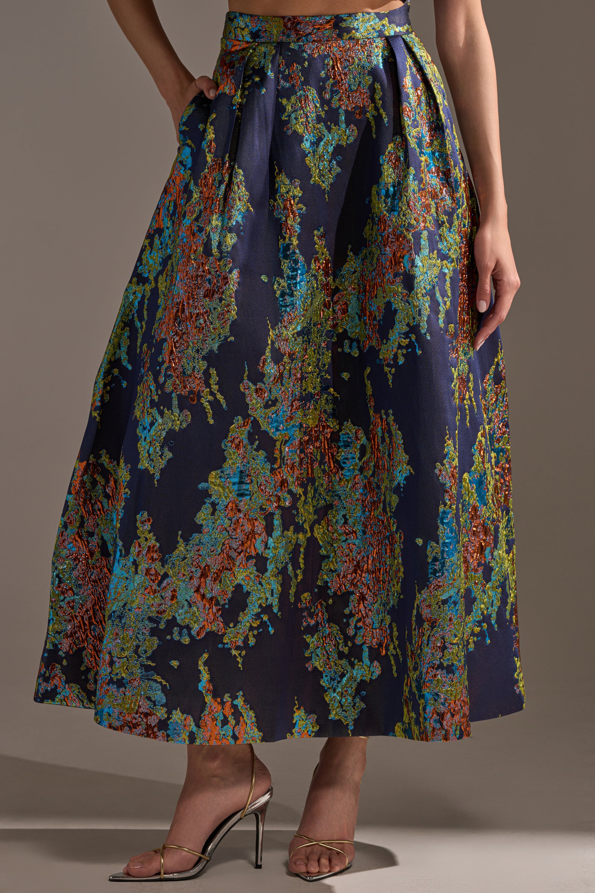 Someone is wearing the IN THE WILD BROCADE MAXI SKIRT, a long dark blue pleated maxi skirt with a vibrant abstract green, orange, and blue pattern, styled with silver high-heeled sandals.