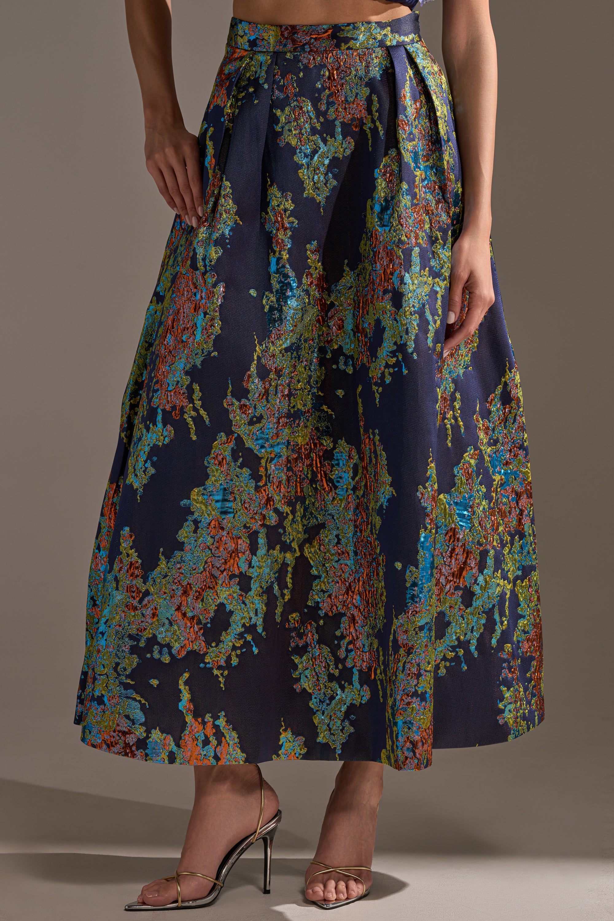 A person wears the IN THE WILD BROCADE MAXI SKIRT, a navy blue high-rise maxi with vibrant abstract patterns in green, orange, and blue, paired with metallic high-heeled sandals. The top is not visible.
