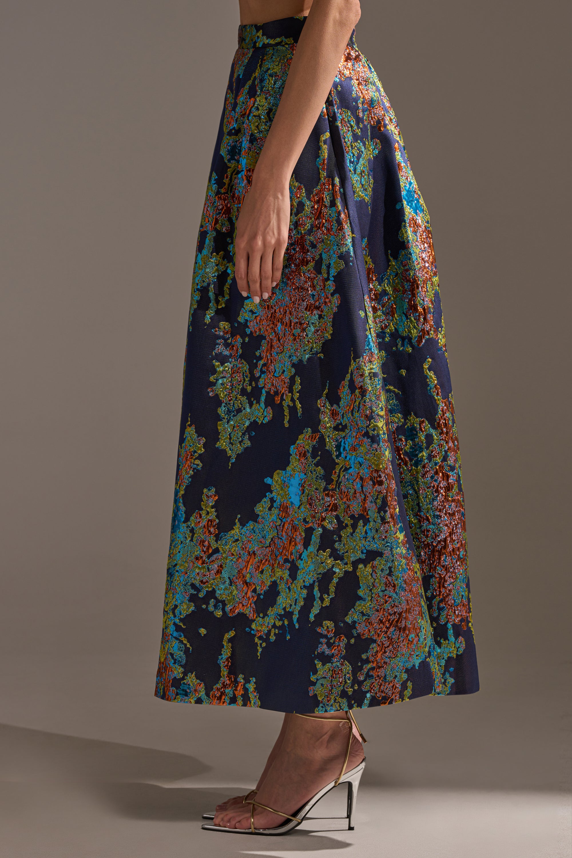 A person stands in profile against a plain background, wearing the IN THE WILD BROCADE MAXI SKIRT with a colorful abstract pattern and silver open-toe heels.