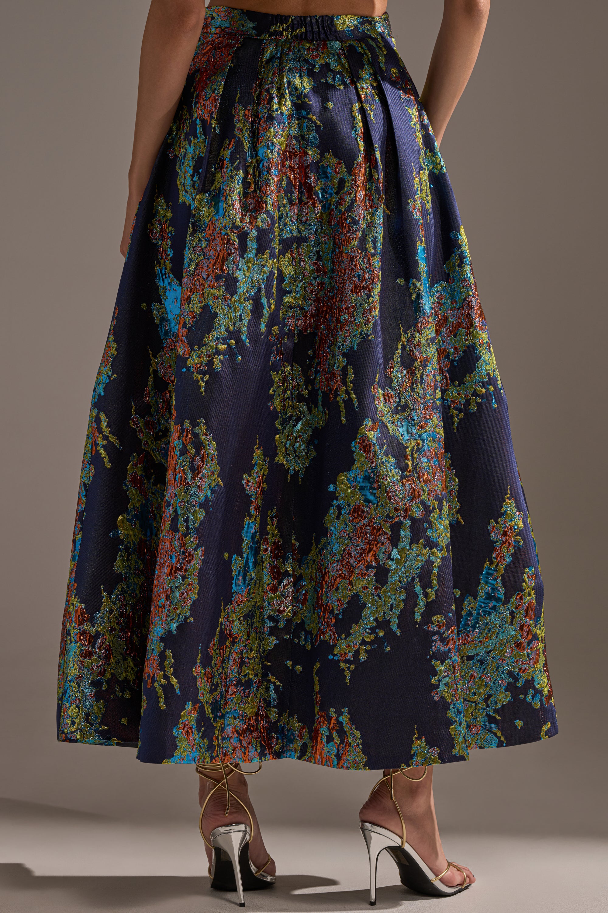 A person wears the IN THE WILD BROCADE MAXI SKIRT, featuring a metallic finish and colorful abstract floral pattern, paired with metallic high-heeled sandals. The photo is taken from behind, showing only the lower half of the body.