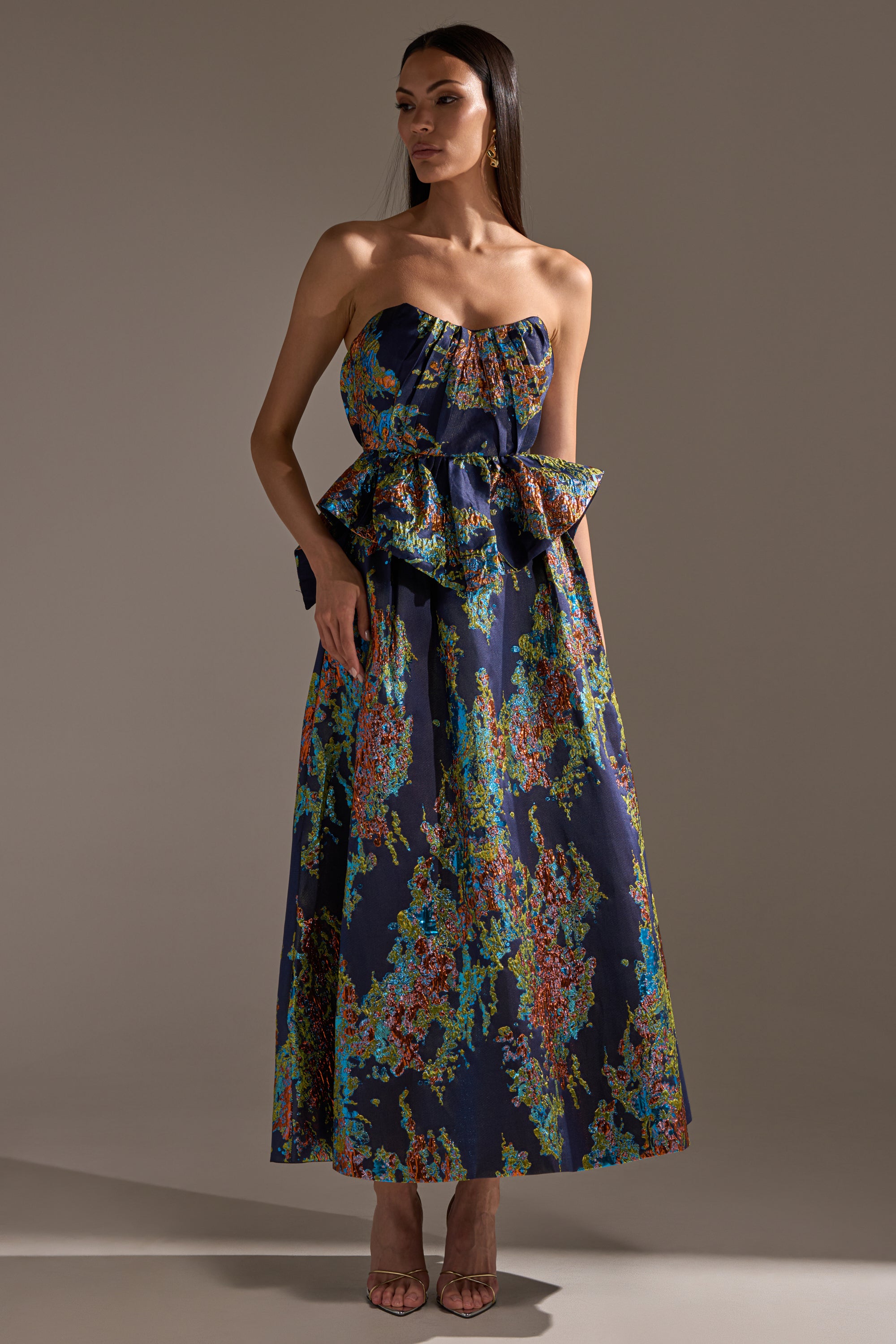 A woman wearing the IN THE WILD BROCADE MAXI SKIRT, featuring a strapless navy blue design with pleats, a colorful abstract floral pattern, and a large bow at the waist, stands in a neutral studio. She has straight hair and open-toe heels.