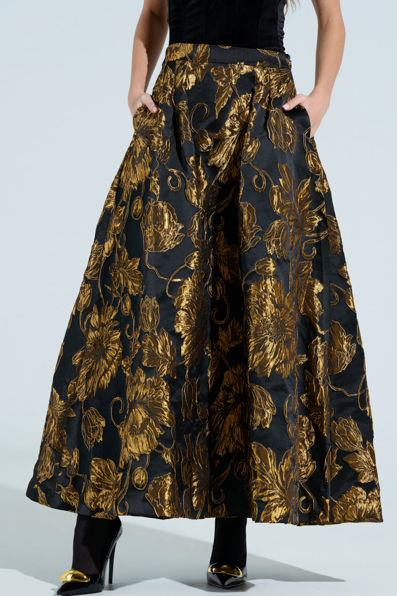 A person wears the stylish IN THE WILD FLORAL BROCADE MAXI SKIRT, with gold patterns on a dark, satin-like fabric, paired elegantly with a black top. The skirt flows to the floor as they casually place their hands in its pockets and complete the look with black shoes.