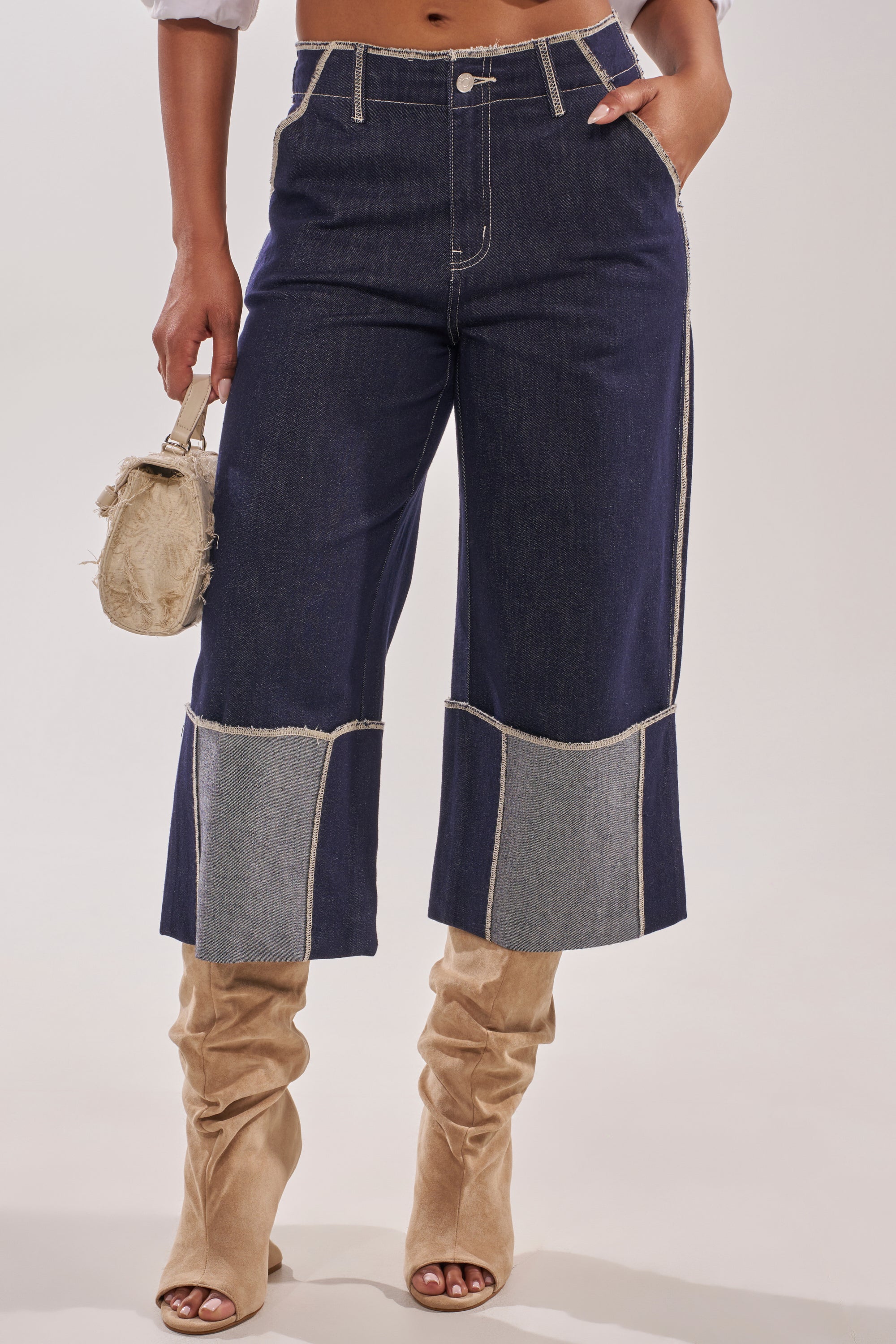 A person wears the LOCKED UP DENIM BERMUDA SHORT, dark blue with light gray patchwork at the hem, paired with beige slouchy heeled boots and a small beige textured handbag. 0