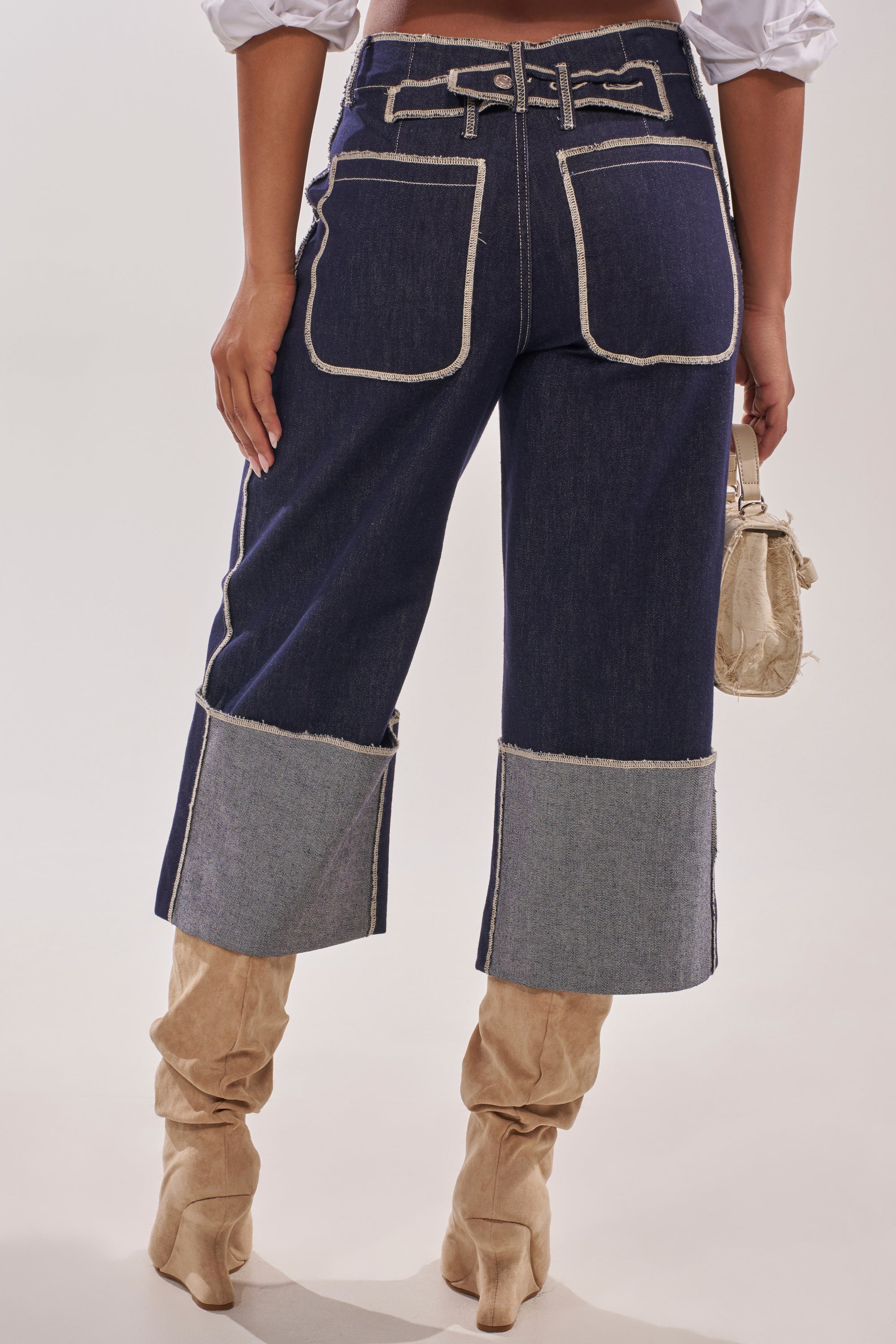A person wearing the LOCKED UP DENIM BERMUDA SHORT—high-waisted with visible white stitching, large back pockets, and contrasting cuffed hems—paired with beige slouchy boots and a small beige handbag. 3