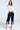 A woman stands confidently wearing the LOCKED UP DENIM BERMUDA SHORT, a high-rise, dark blue bermuda short with light denim cuffs. She pairs them with an off-the-shoulder white blouse and pointed-toe heels, her hands in her pockets.
