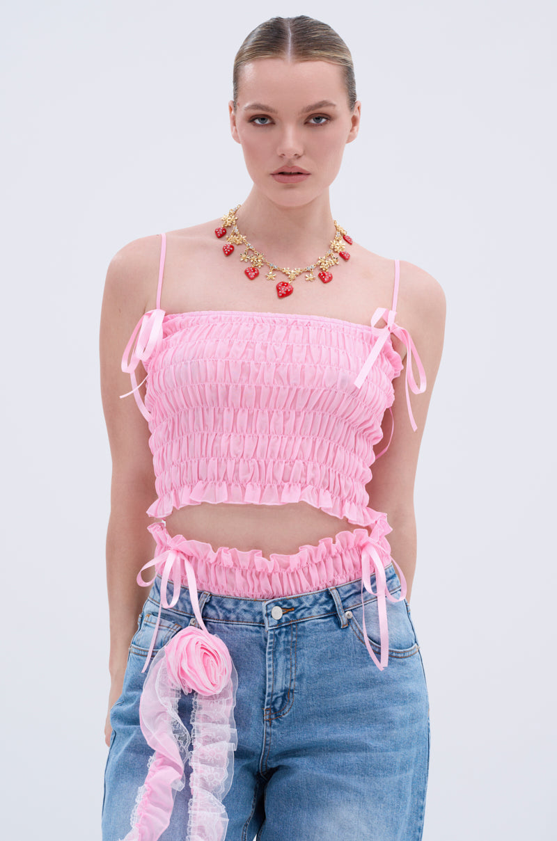 A woman stands against a plain light background, hair slicked back, wearing the CALL ME RUCHED BOW TANK in pink with ribbon ties, blue jeans, and a gold necklace featuring red heart-shaped charms.