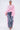 A woman stands against a plain light background wearing FLOWER GIRLIE APPLIQUE DENIM JEANS with pink ruffles, a loose light pink top, pink heels, gold hoop earrings, and a gold clutch.