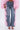 A person wears FLOWER GIRLIE APPLIQUE DENIM JEANS, featuring wide legs and pink ruffle appliqués on the outer seams, styled with a pink textured top, pink strappy heels, and a gold clutch.