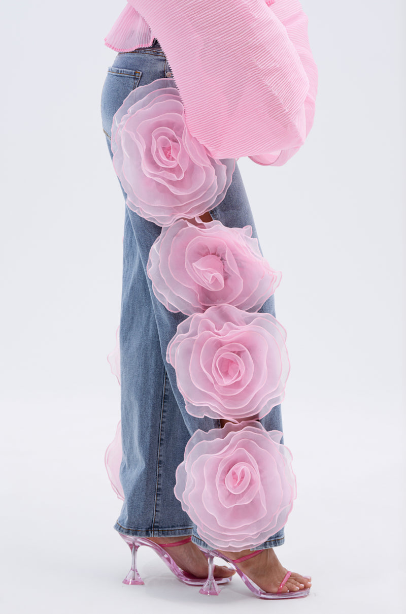 A person wears FLOWER GIRLIE APPLIQUE DENIM JEANS, featuring large layered pink fabric flowers down the leg, styled with a pink pleated top and pink high-heeled sandals against a plain white background.