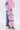 A person wears FLOWER GIRLIE APPLIQUE DENIM JEANS, featuring large layered pink fabric flowers down the leg, styled with a pink pleated top and pink high-heeled sandals against a plain white background.