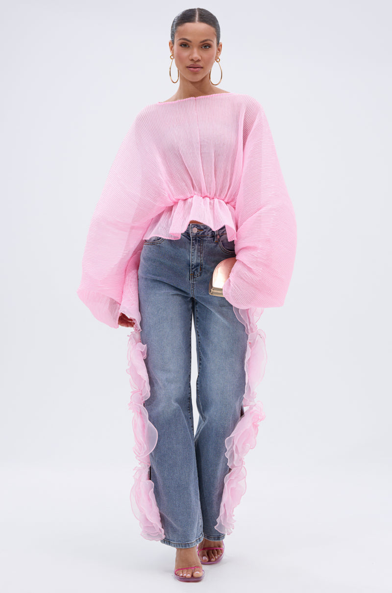 A woman stands facing forward in FLOWER GIRLIE APPLIQUE DENIM JEANS with pink ruffle sides, a sheer oversized pink top, hoop earrings, pink heeled sandals, and holding a small purse against a plain background.