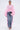 A woman stands facing forward in FLOWER GIRLIE APPLIQUE DENIM JEANS with pink ruffle sides, a sheer oversized pink top, hoop earrings, pink heeled sandals, and holding a small purse against a plain background.