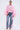 A woman stands against a plain background wearing FLOWER GIRLIE APPLIQUE DENIM JEANS paired with a sheer, oversized pink top, featuring billowy sleeves and flowing sides, along with pink high-heeled sandals.