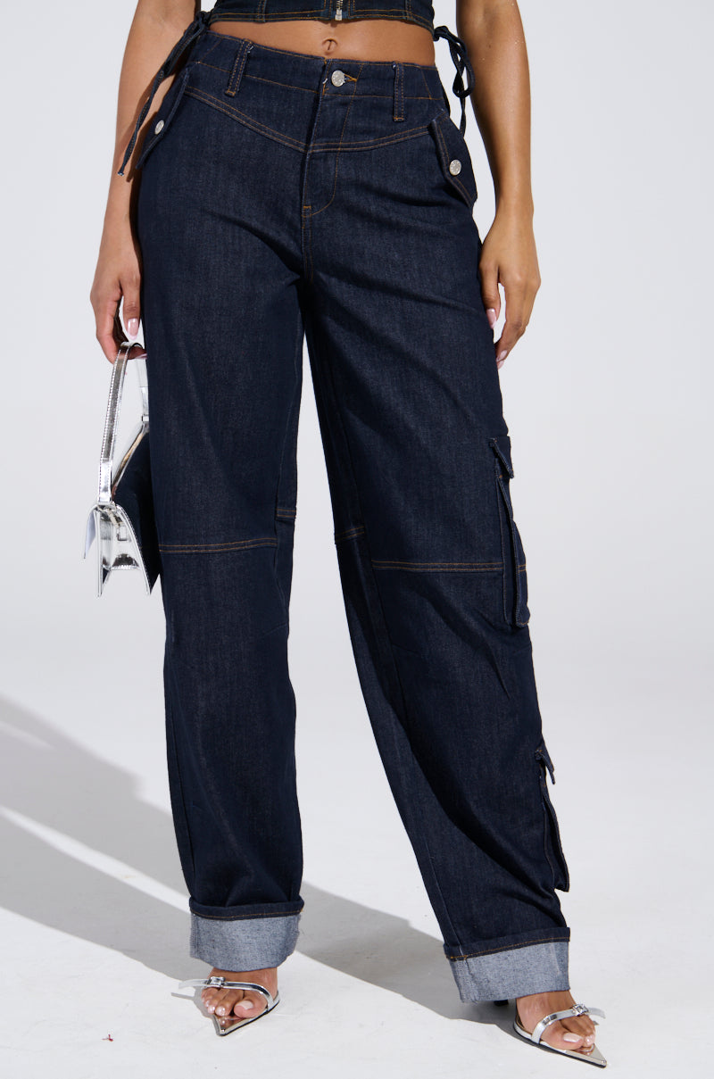 A person wears the DENIM FOR DAYS DENIM PANT with a high-rise fit and cargo pockets, styled with a black crop top, silver heels, and a metallic handbag. The background is plain and light-colored.