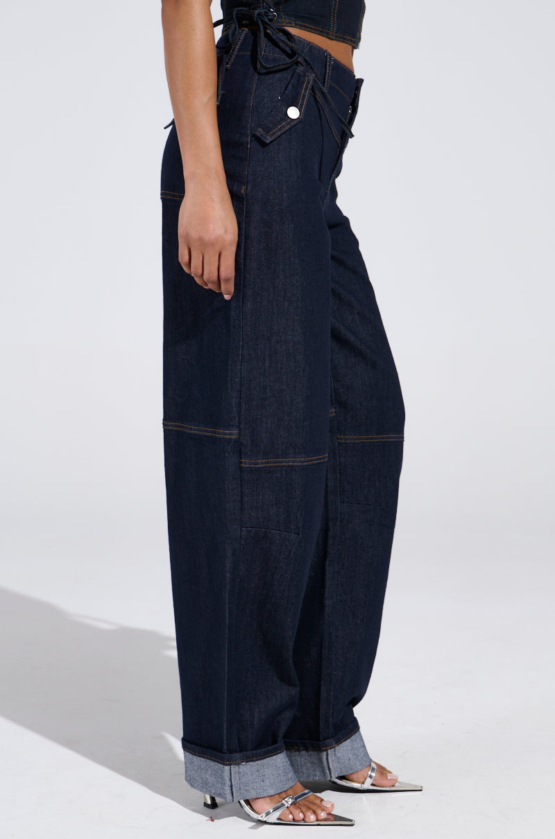 Someone models the DENIM FOR DAYS DENIM PANT—dark-wash, high-rise wide-leg jeans with cuffed hems—paired with white heeled sandals, shown in a side profile against a plain light background.
