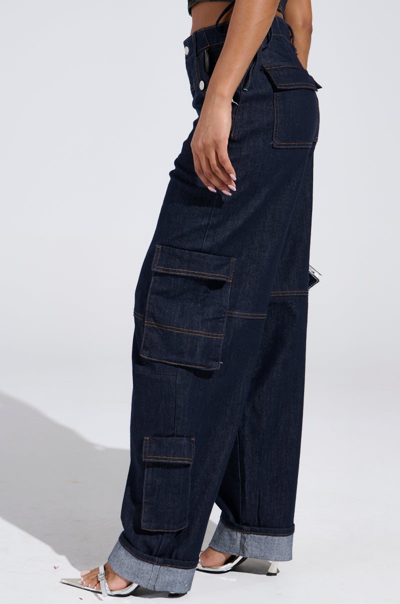 A person wears the DENIM FOR DAYS DENIM PANT featuring a high-rise fit, dark wash, wide legs, and cargo pockets. The jeans are cuffed at the ankles and paired with white heeled sandals for a chic, modern look.