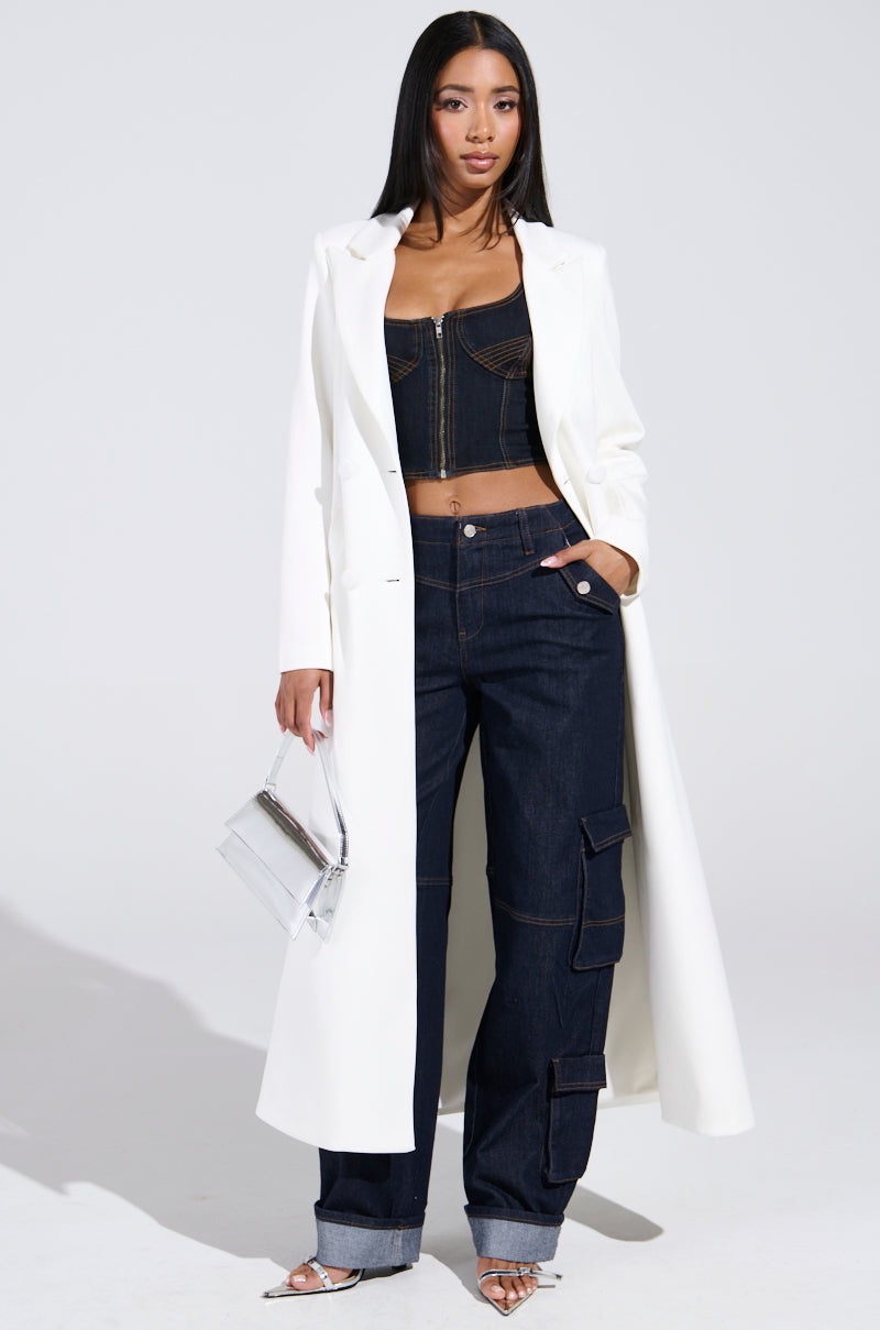 A woman stands confidently in a long white coat over a black zip-up bustier top, paired with DENIM FOR DAYS DENIM PANT featuring cargo-style pockets, silver heels, and a small metallic silver handbag. Her straight hair falls loose.