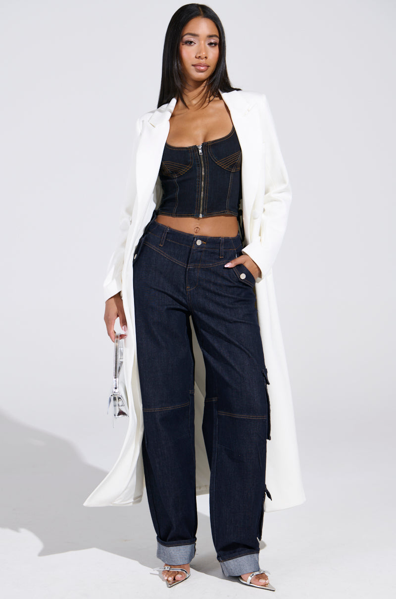 A woman stands confidently in a long white coat over a black zip-up crop top and DENIM FOR DAYS DENIM PANT with rolled cuffs, holding a small clear handbag.