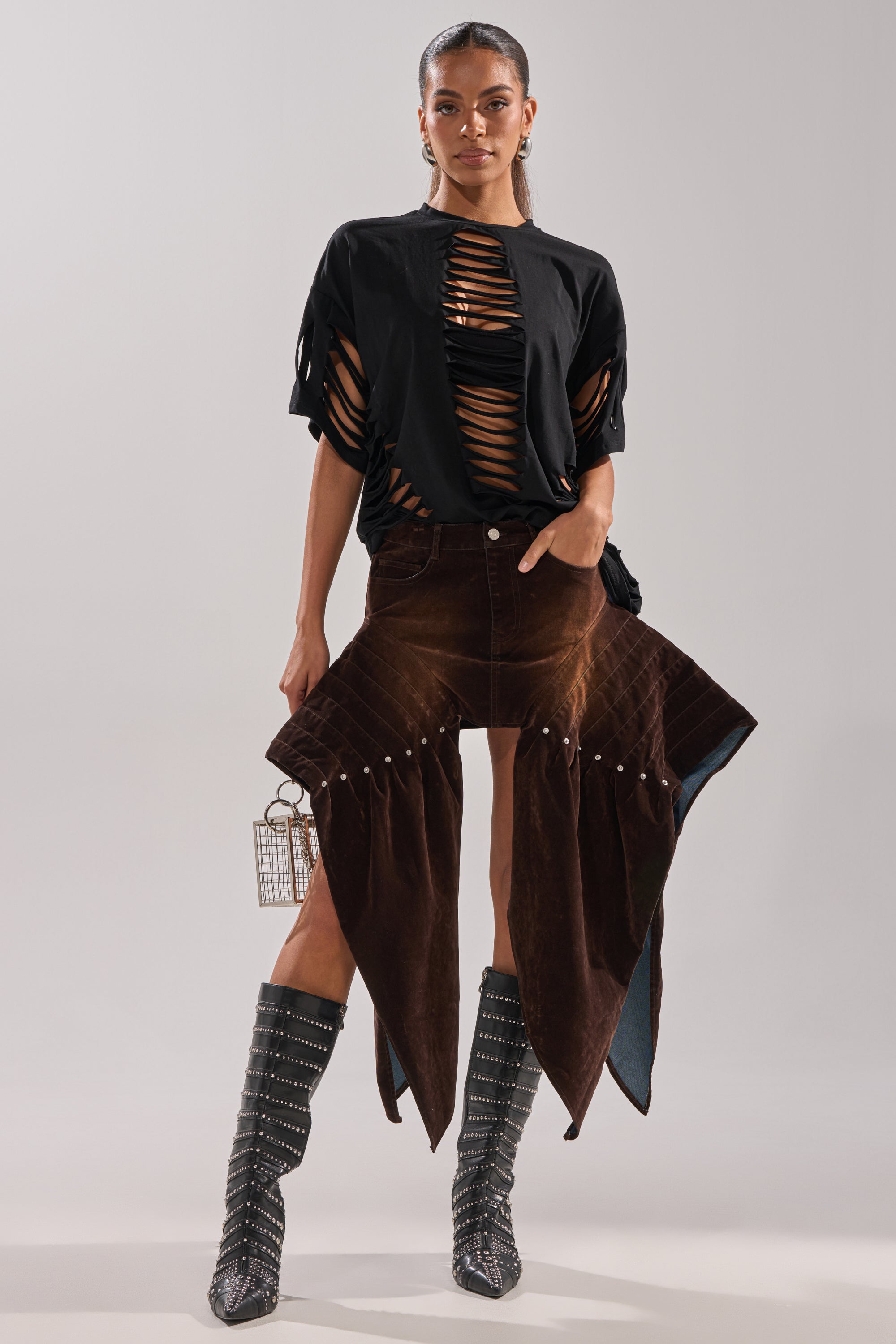 A woman stands against a plain background wearing a black, shredded cut-out top, patterned knee-high boots, holding a small box handbag, and the ALL FOR IN VELVET DENIM MINI SKIRT with high-rise pointed panels.
