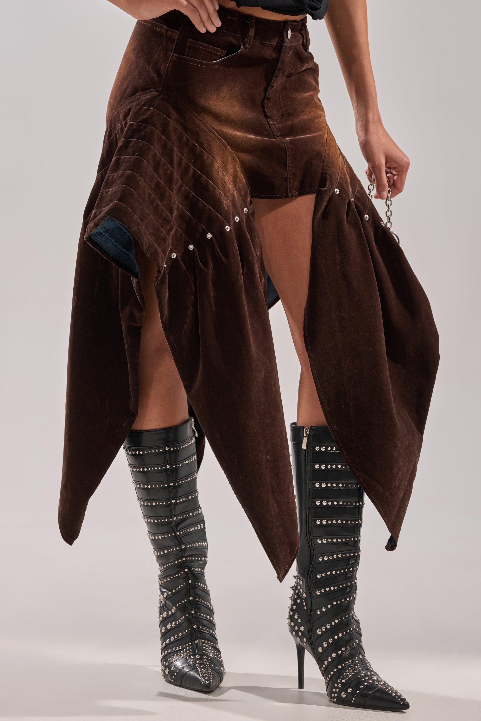 Wearing the ALL FOR IN VELVET DENIM MINI SKIRT in brown-wash with pointed panels and metal studs, styled with black high-heeled boots featuring silver studs and finished with a small chain accessory.