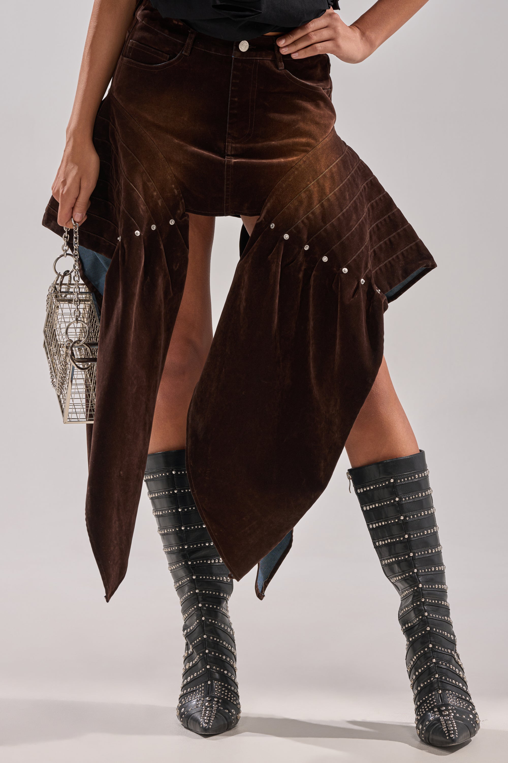 A person showcasing only their lower half wears the ALL FOR IN VELVET DENIM MINI SKIRT in brown velvet denim with silver studs, styled with studded black knee-high boots and a metallic wire handbag.