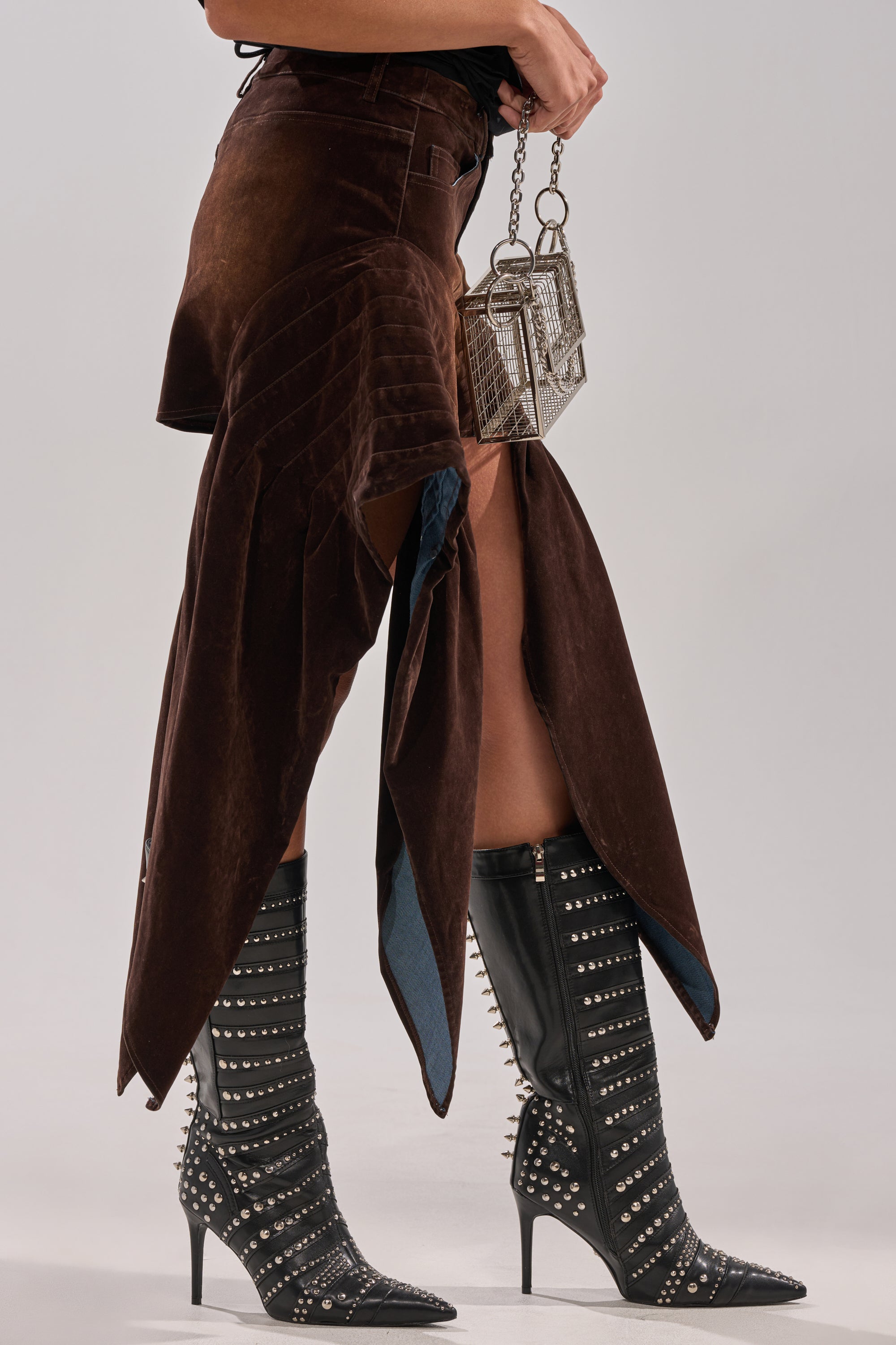 A person holds a small silver handbag, wearing the ALL FOR IN VELVET DENIM MINI SKIRT and black knee-high boots with pointed toes, studs, and spikes.
