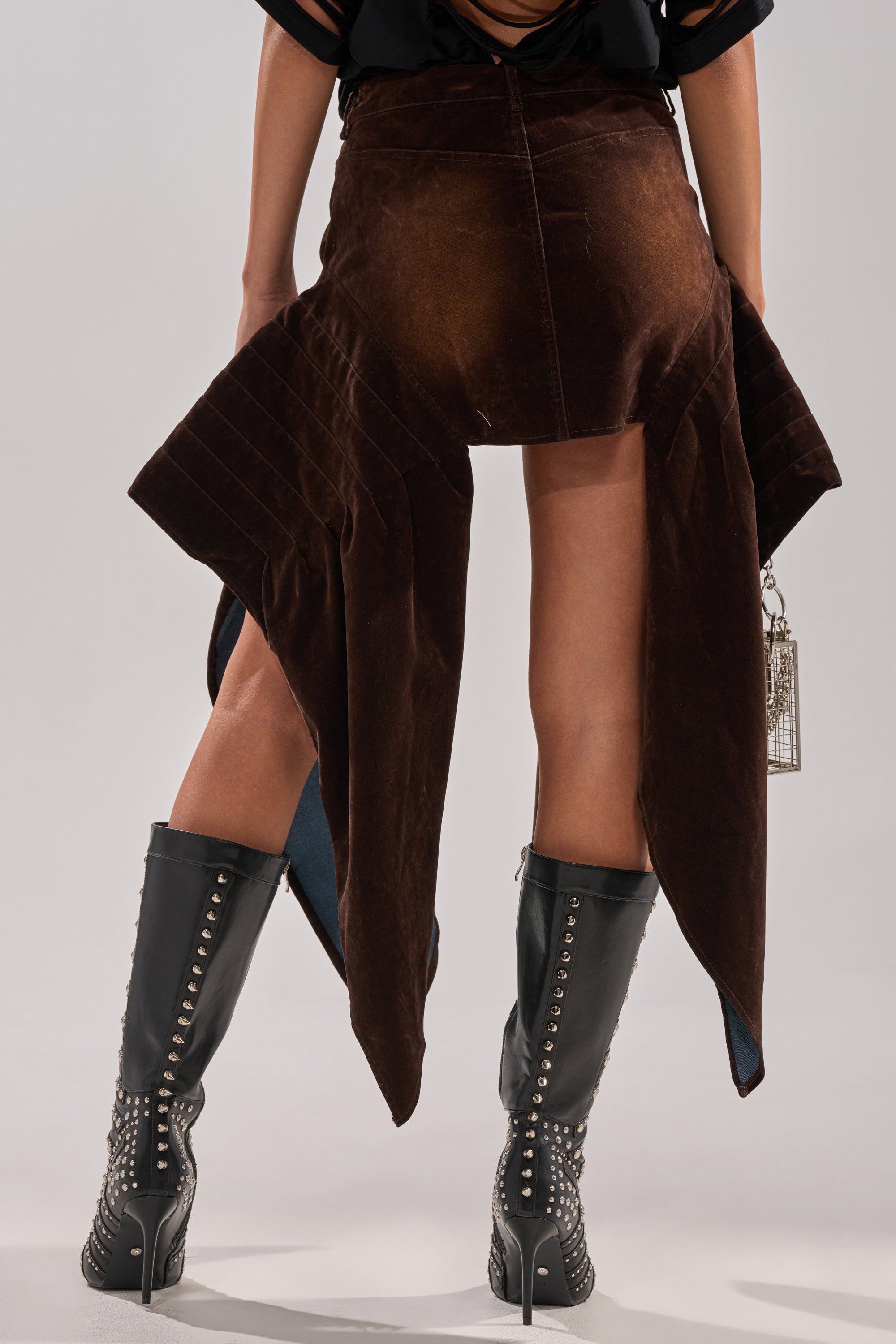 A person stands facing away, wearing the ALL FOR IN VELVET DENIM MINI SKIRT in dark brown with draped panels, paired with black knee-high boots featuring silver studs and holding a small metallic rectangular purse.
