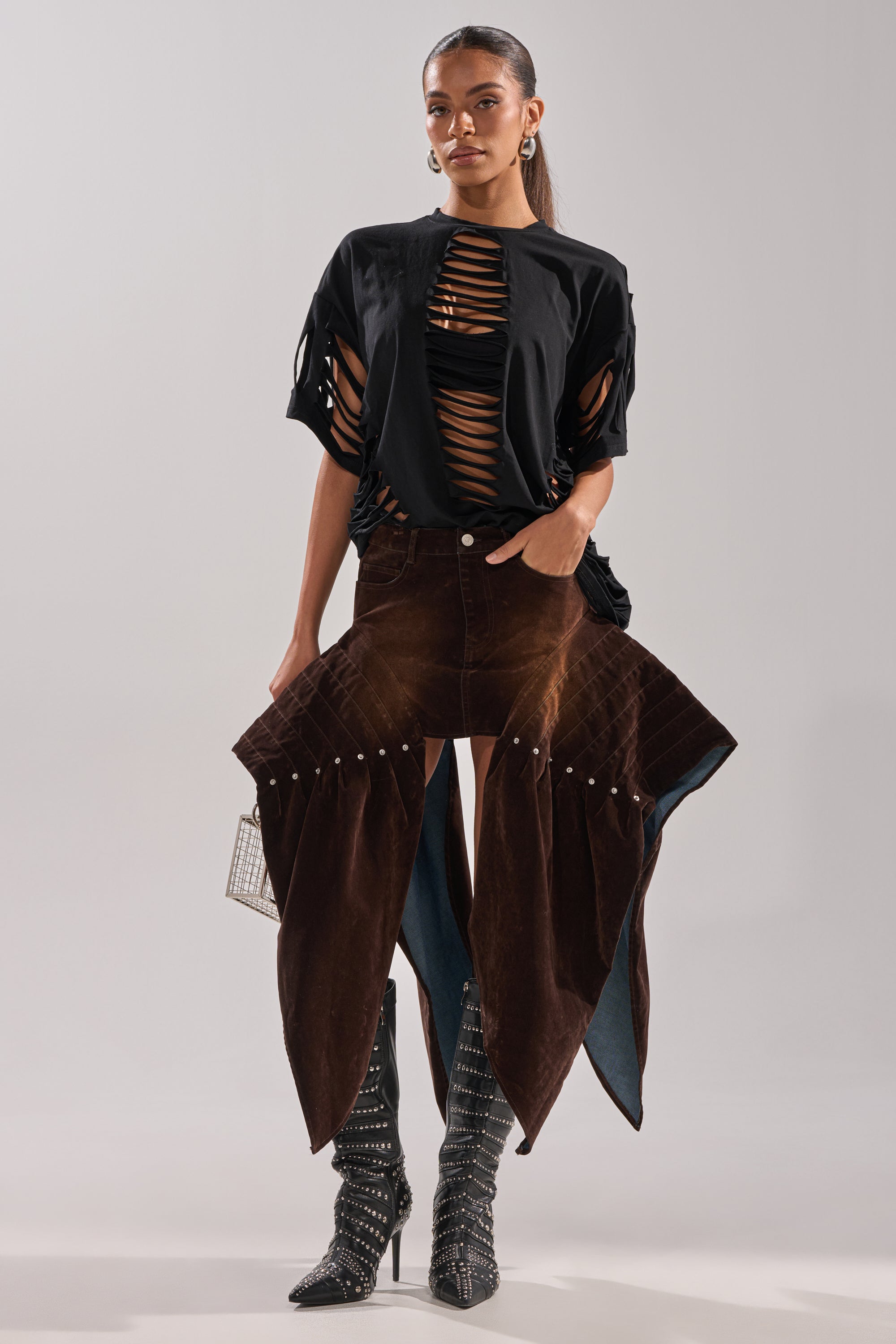 A woman models the ALL FOR IN VELVET DENIM MINI SKIRT in a brown wash with pointed flaps, paired with a black shredded top, matching bralette, patterned knee-high boots, hoop earrings, and a small square handbag against a plain background.