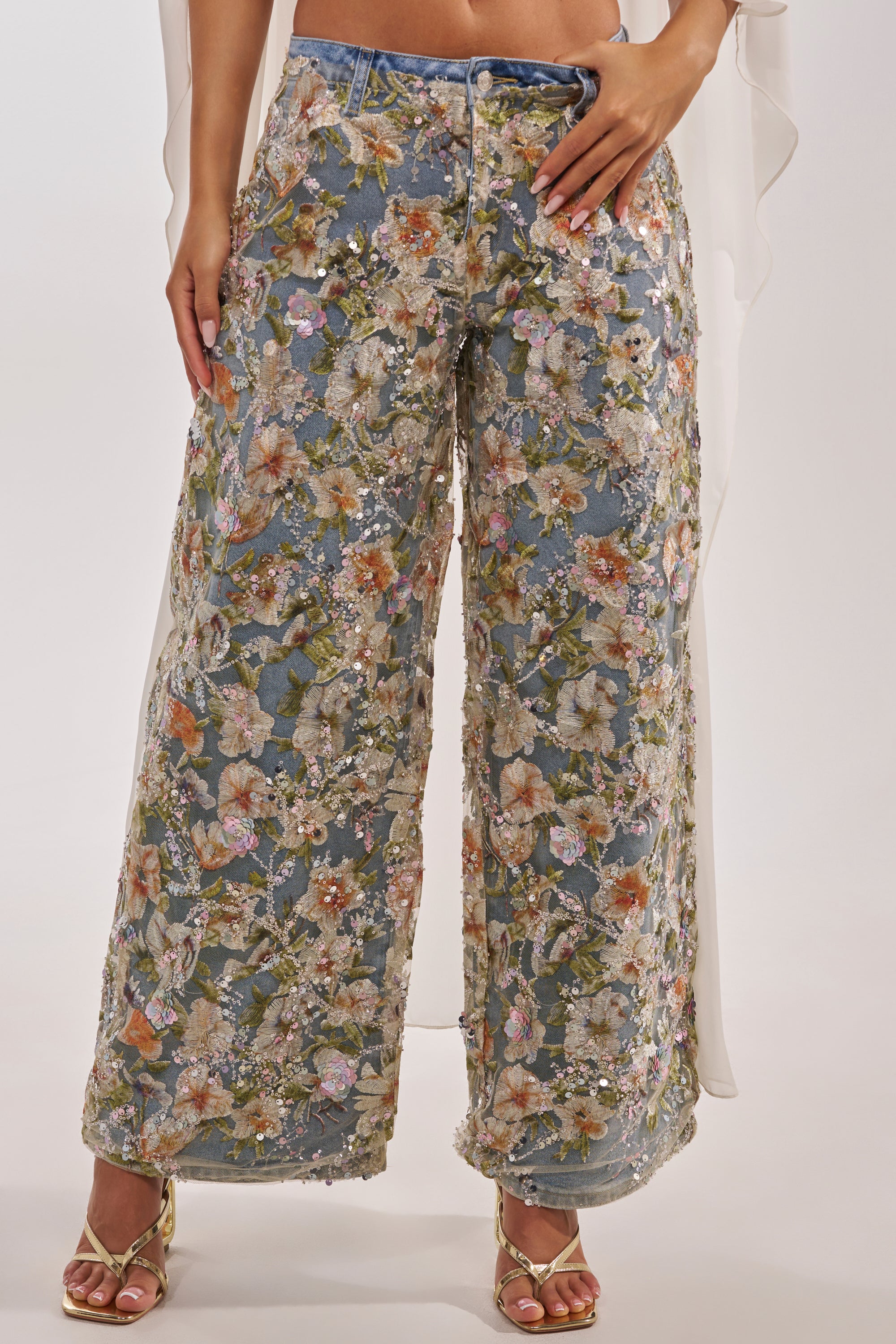 Wearing the IRIS RELAXED DENIM WITH FLORAL OVERLAY, featuring colorful embroidery and sequins, paired with gold open-toe heels, a person stands against a plain background. 0