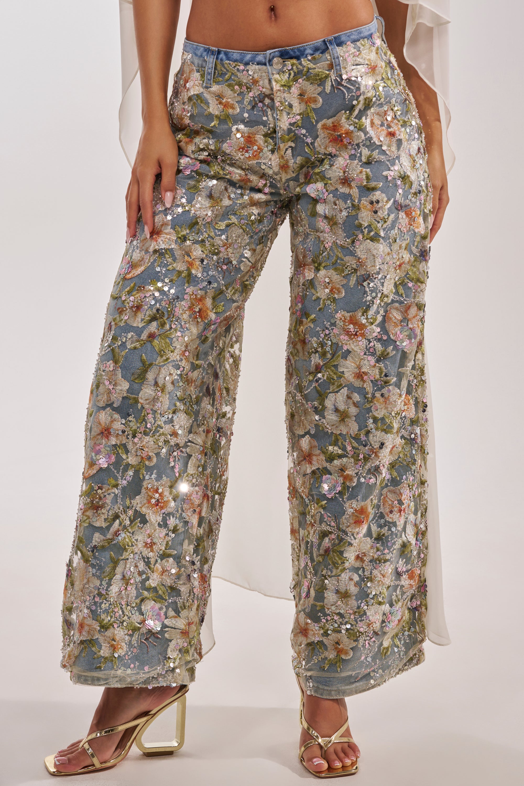 Someone wearing IRIS RELAXED DENIM WITH FLORAL OVERLAY and strappy gold high heels stands against a plain background. 1