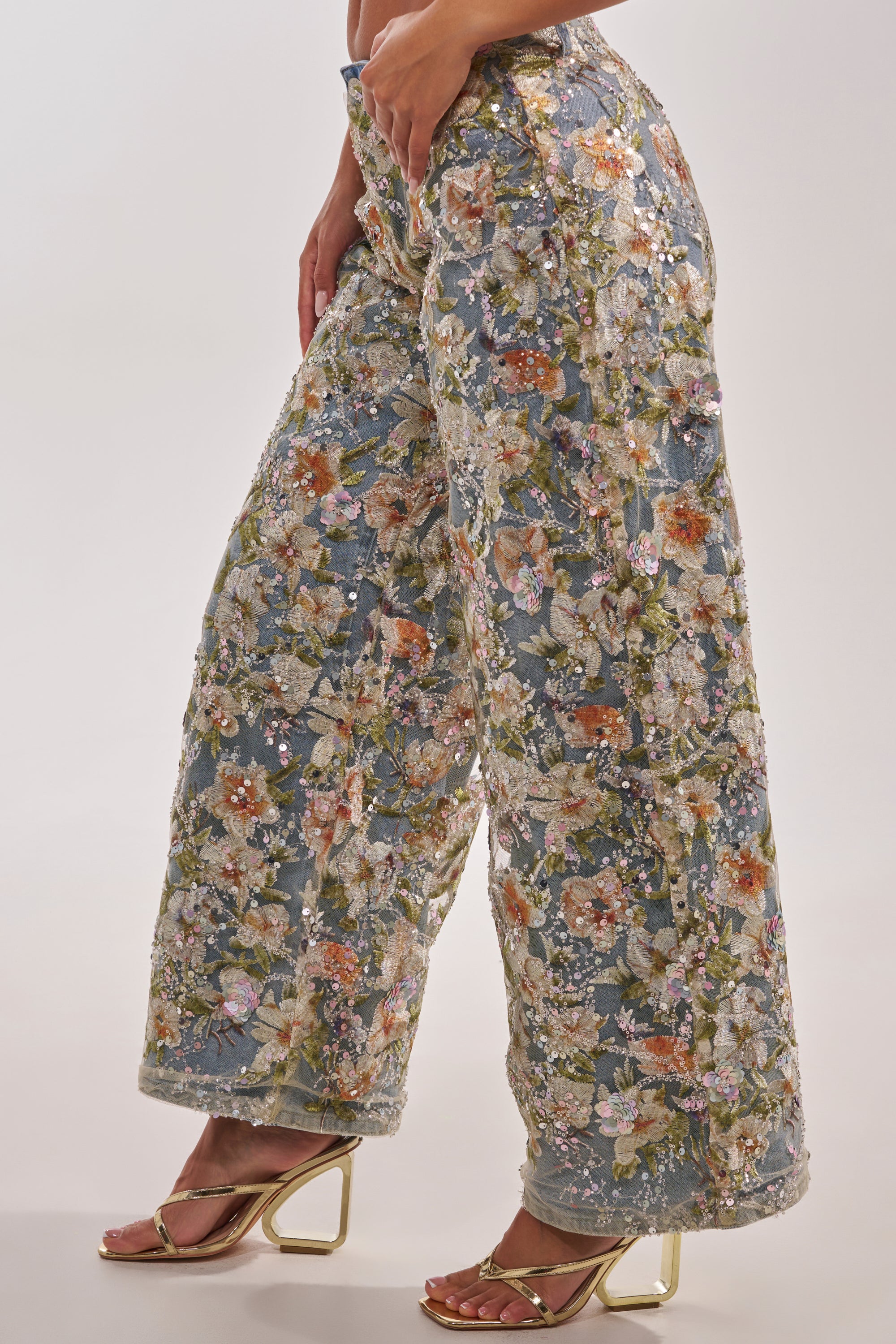 Someone in the IRIS RELAXED DENIM WITH FLORAL OVERLAY—featuring an intricate multicolor floral design—paired with metallic heeled sandals, stands against a plain, light background. 2