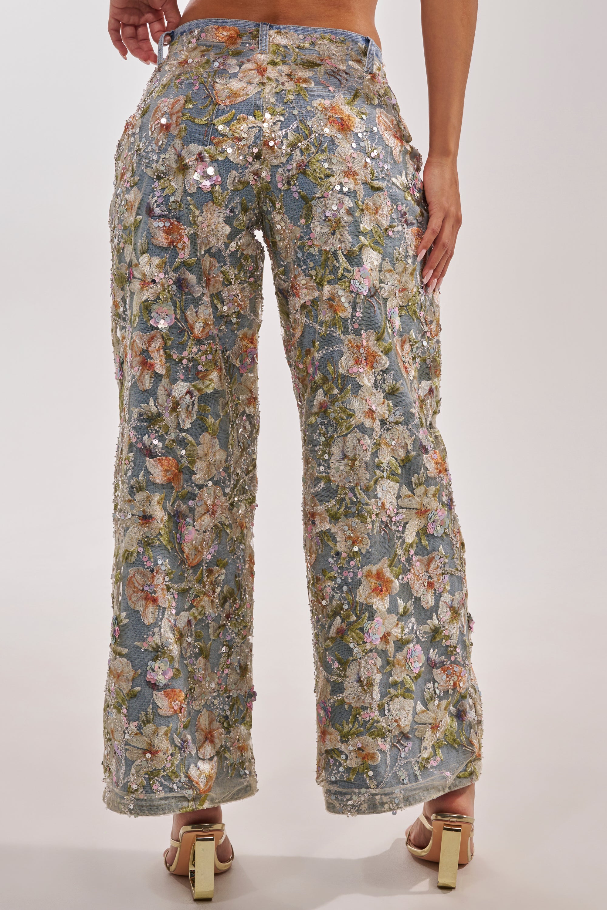 A person stands wearing the IRIS RELAXED DENIM WITH FLORAL OVERLAY, featuring high-rise wide-leg jeans adorned with colorful embroidered florals and sequins. The back view emphasizes the detailed embellishments. 3